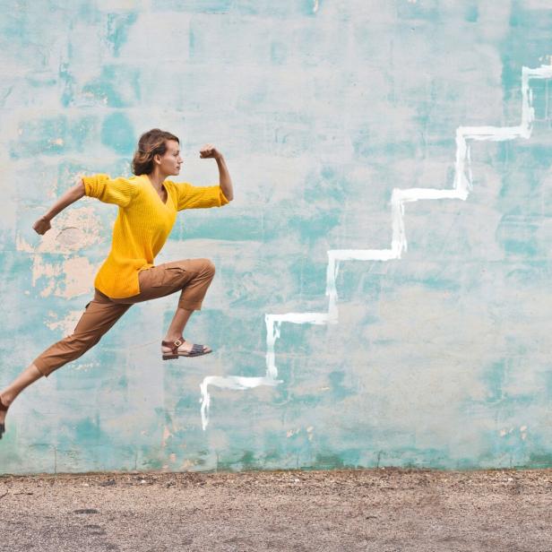 Eine Frau springt dynamisch vor einer Wand mit aufgemalten weißen Treppenstufen nach oben.