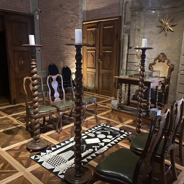 Ein Raum mit Stühlen im Kreis um einen Teppich, flankiert von Kerzenständern.