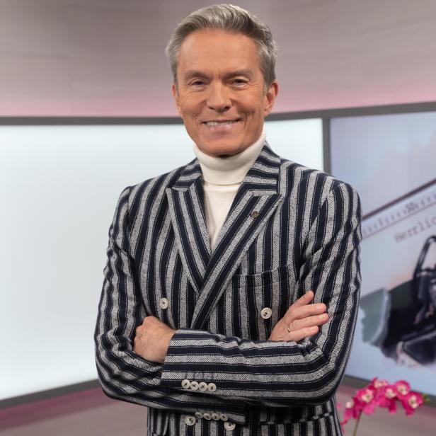 Ein lächelnder Mann mit gestreiftem Sakko und Rollkragenpullover steht vor einem Bildschirm.