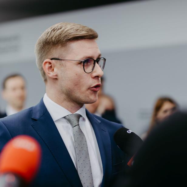 Ein Mann mit Brille und Anzug spricht in Mikrofone vor unscharfem Hintergrund.