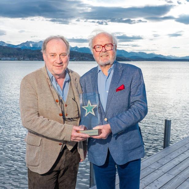 Zwei Männer halten eine Auszeichnung vor einem See und Bergen.