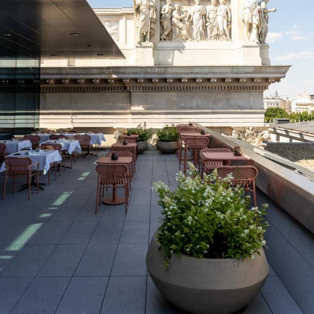 Ein Dachterrassen-Restaurant mit Tischen und Stühlen befindet sich unterhalb eines Gebäudes mit Skulpturen.