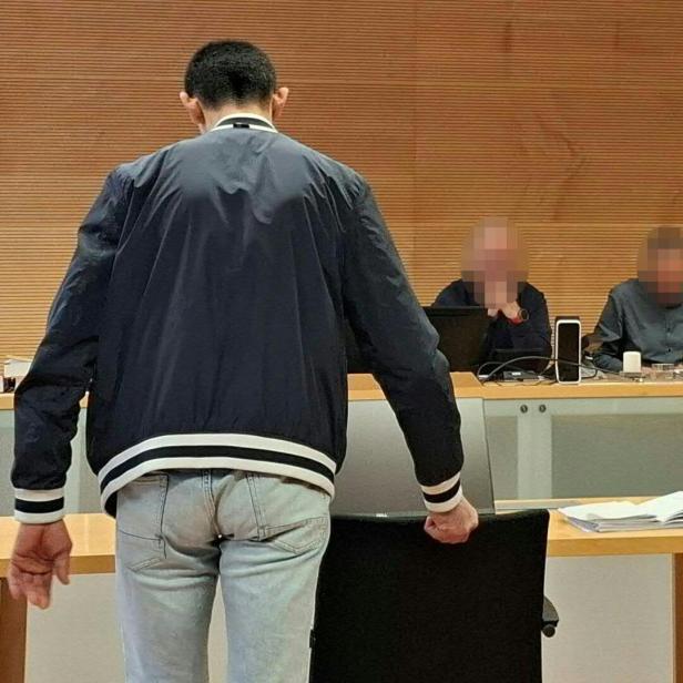 Ein Mann steht vor einem Tisch mit zwei Personen in einem Gerichtssaal.