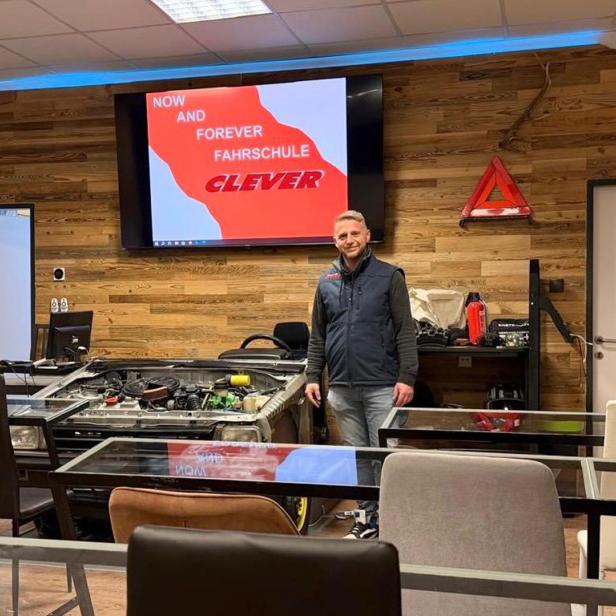 Ein Fahrlehrer der Fahrschule Clever Graz steht in einem modernen Unterrichtsraum mit Motorenmodell und Präsentationsbildschirm.