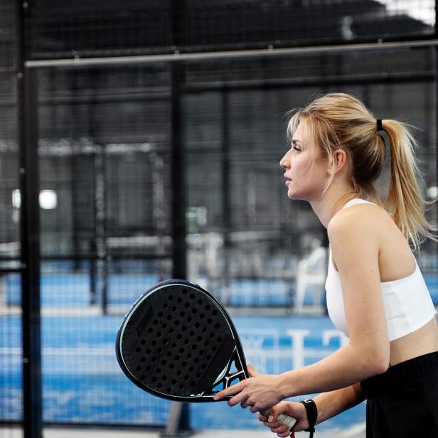 Junge Frau mit blondem Pferdeschwanz hält einen Padel-Schläger und steht konzentriert auf einem blauen Padelplatz.