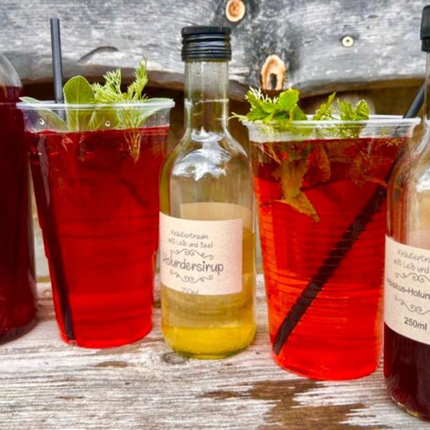 Verschiedene Flaschen und Becher mit roten Kräutergetränken und frischen Kräutern auf Holztisch.