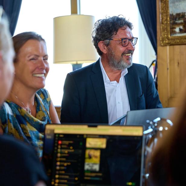 Mehrere Personen sitzen lachend an einem Tisch mit Laptops in einem hellen Raum.