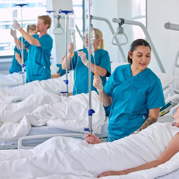 Pflegekräfte betreuen mehrere Patientinnen und Patienten in Krankenhausbetten und legen Infusionen an.