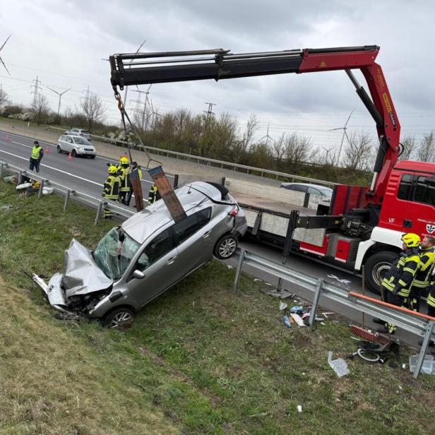 Ein silbernes Auto mit starkem Frontschaden wird von Feuerwehrleuten und einem Kran von einer Böschung neben einer Straße geborgen.