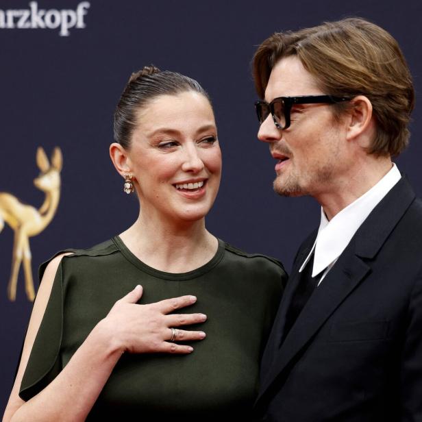 Alexandra Maria Lara und Sam Riley elegant gekleidet vor grauem Hintergrund.