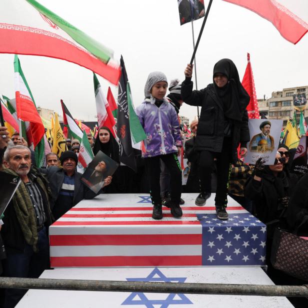 Zwei Kinder stehen auf einer Plattform mit US-Flagge und Davidstern, umgeben von Menschen mit iranischen Fahnen und Porträts.