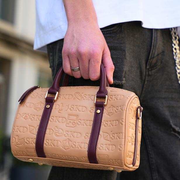 Beige SKD-Handtasche mit geprägtem Logo und braunen Henkeln wird von einer Person getragen.