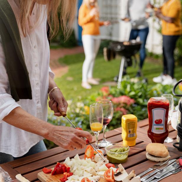 Frau bereitet Gemüse am Tisch vor, im Hintergrund grillen mehrere Personen im Garten.