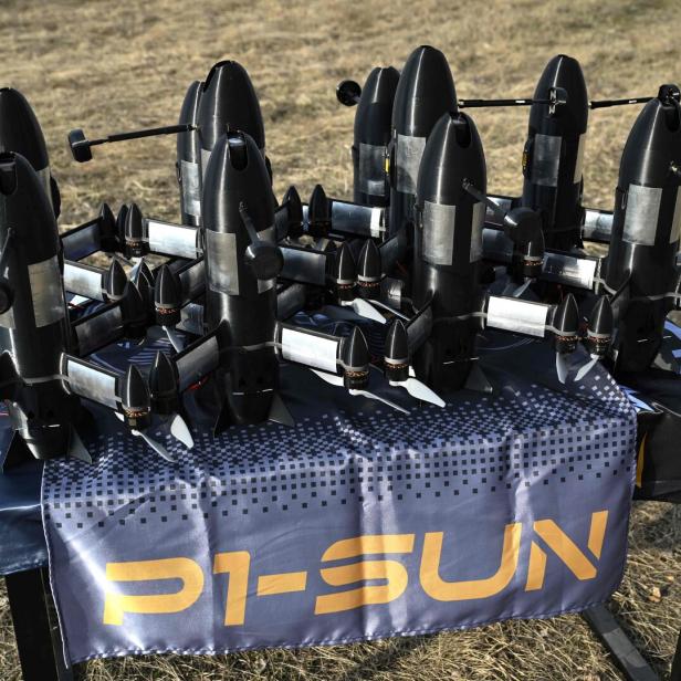 Mehrere schwarze Drohnen mit Propellern stehen auf einem Tisch im Freien auf einer blauen Decke mit gelber Aufschrift.