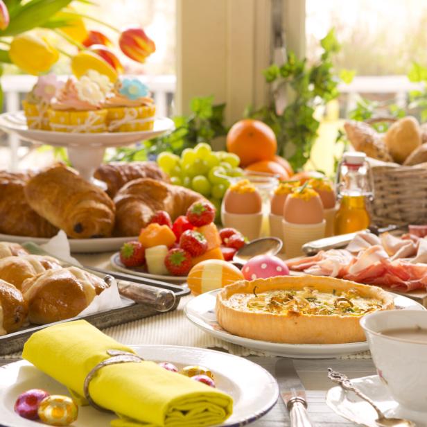 Ein reichhaltiger Brunch mit Gebäck, Quiche, Obst, Eiern und Tulpen steht auf einem Tisch.