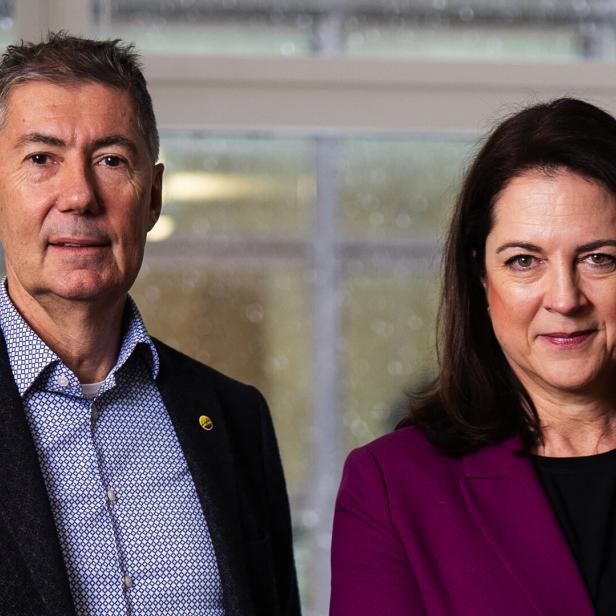 Ein Mann im Sakko und eine Frau im lila Blazer stehen nebeneinander vor einem Fenster.