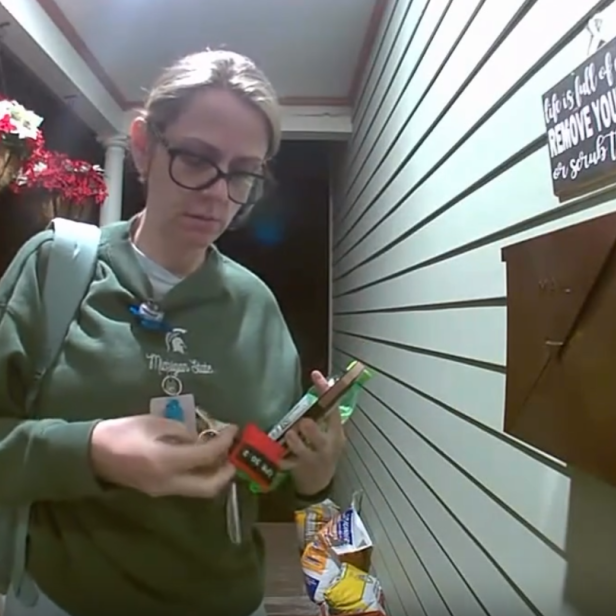 Eine erschöpfte Frau steht mit Ausweis und Schlüsseln vor einer Haustür im Eingangsbereich.