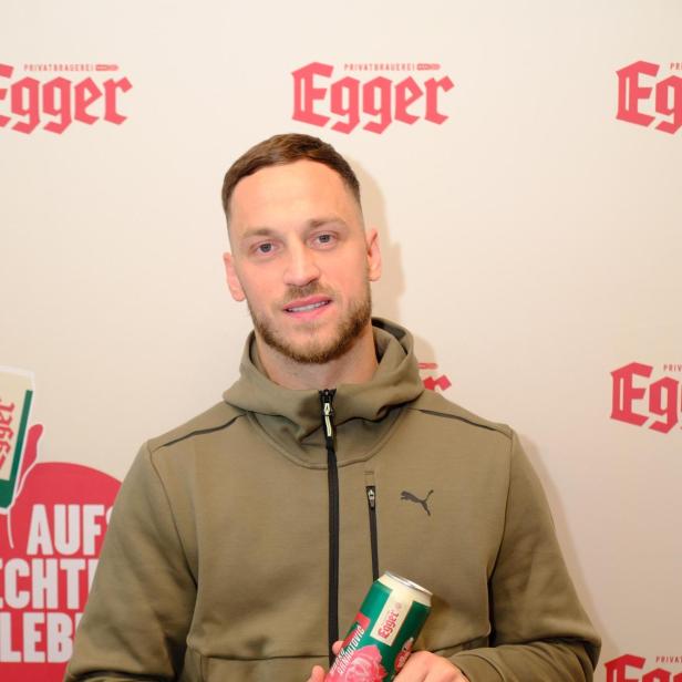 Ein Mann in olivgrüner Jacke hält eine Getränkedose mit rotem Etikett vor einer Wand mit Egger-Logos.