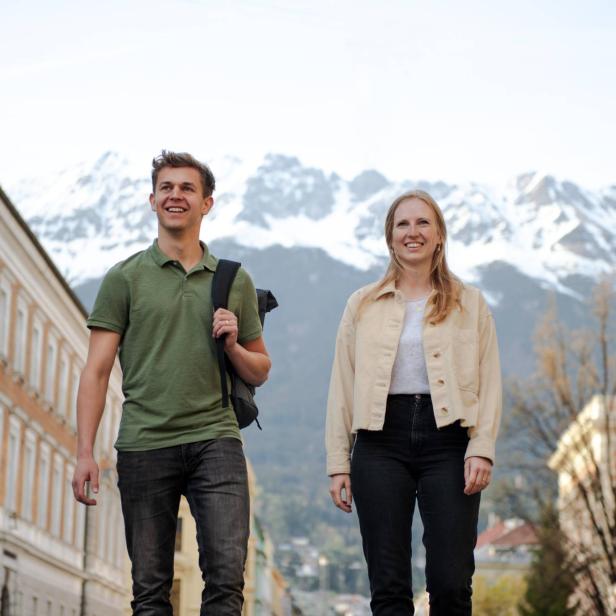 Zwei junge Menschen gehen lächelnd durch eine Stadt mit schneebedeckten Bergen im Hintergrund.
