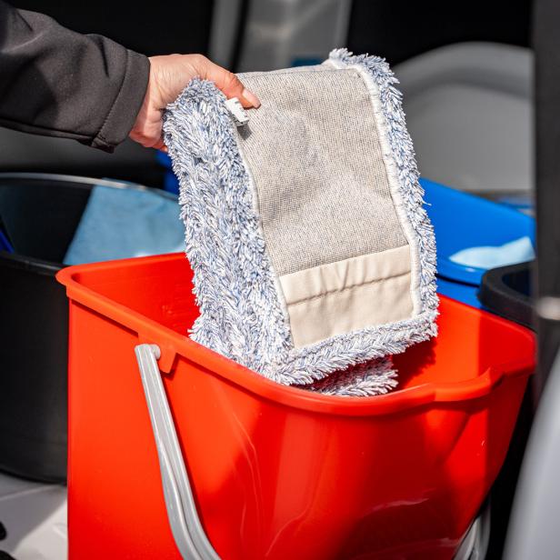 Hand legt einen Wischmopp in einen roten Putzeimer, daneben stehen weitere Reinigungseimer.