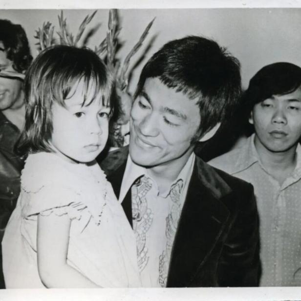 Bruce Lee hält ein kleines Mädchen im Arm, während andere Personen im Hintergrund stehen.