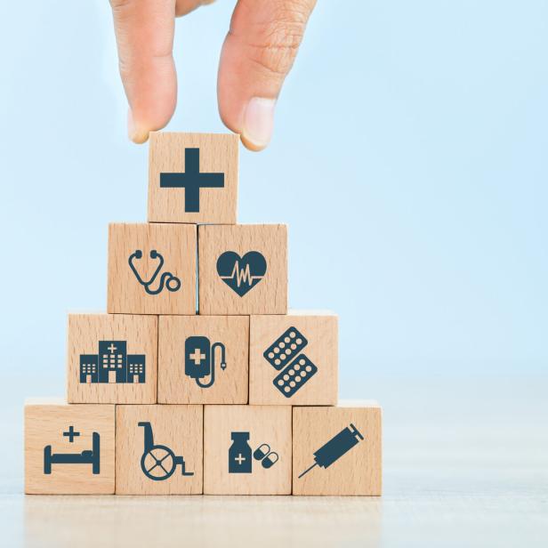Das Foto zeigt zu einem Turm aufgestapelte Holzwürfel mit kleinen graphischen Zeichen auf der Vorderseite, die symbolhaft für das Gesundheitssystem stehen wie etwa ein Stethoskop, eine EKG-Linie oder eine Spritze.