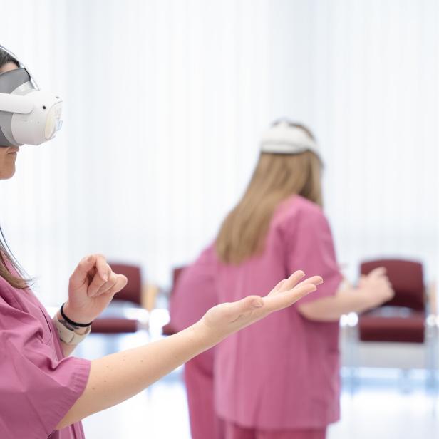 Zwei Personen in rosa Kitteln tragen Virtual-Reality-Headsets und führen gestikulierende Bewegungen in einem hellen Raum aus.