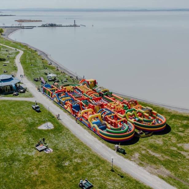 Großer bunter Hüpfburgen-Parcours am Ufer eines Sees, umgeben von Wiese, Wegen und Bäumen mit Menschen.