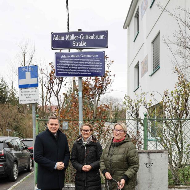 Drei Personen und ein Hund stehen in der Adam-Müller-Guttenbrunn-Straße vor einem Straßenschild mit Informationstafel.