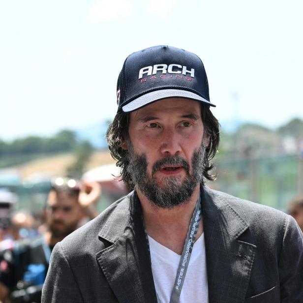Keanu Reeves mit Bart und Kappe "Arch Racing" trägt ein Jackett.