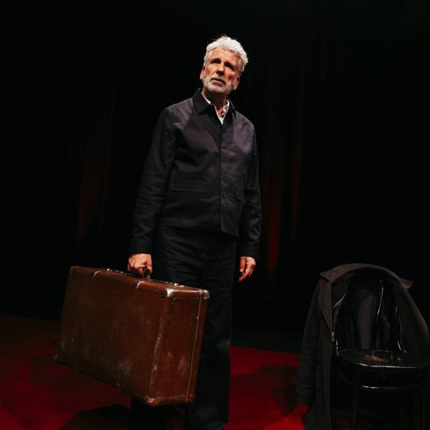 Dirk Stermann hält einen alten Koffer vor schwarzem Hintergrund.