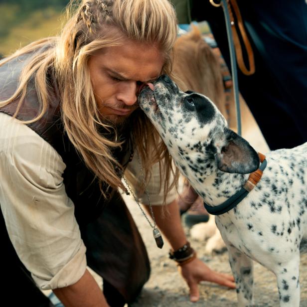 Ein Mann mit langen blonden Haaren wird von einem schwarz-weiß gefleckten Hund im Gesicht abgeschleckt.