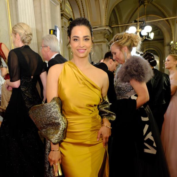 Amira Aly in einem goldenen, schulterfreien Kleid posiert auf einem roten Teppich.