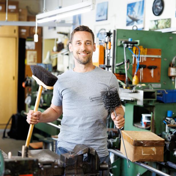 Ein Mann steht in einer Werkstatt und hält einen Besen und eine Bürste in den Händen, umgeben von Maschinen und Werkzeugen.