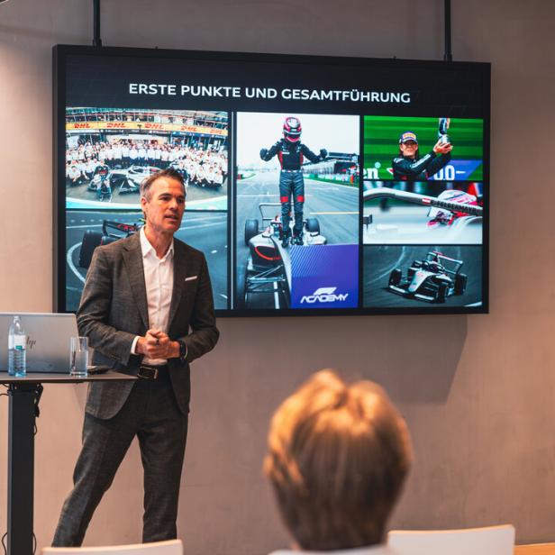 Ein Mann im Anzug hält einen Vortrag vor einer Leinwand mit Motorsport-Bildern und der Überschrift „Erste Punkte und Gesamtführung“.