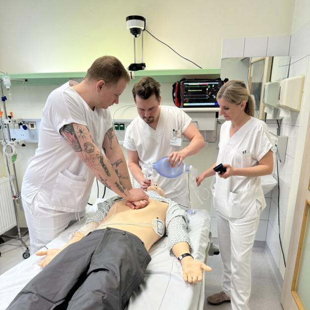Drei Personen in weißer Kleidung üben an einer Reanimationspuppe Herz-Lungen-Wiederbelebung.