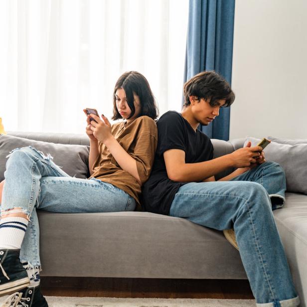 Zwei Jugendliche sitzen Rücken an Rücken auf einem Sofa und schauen konzentriert auf ihre Smartphones.