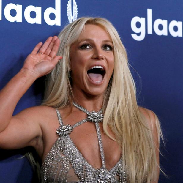 Britney Spears mit erhobener Hand und offenem Mund vor einem GLAAD-Hintergrund.