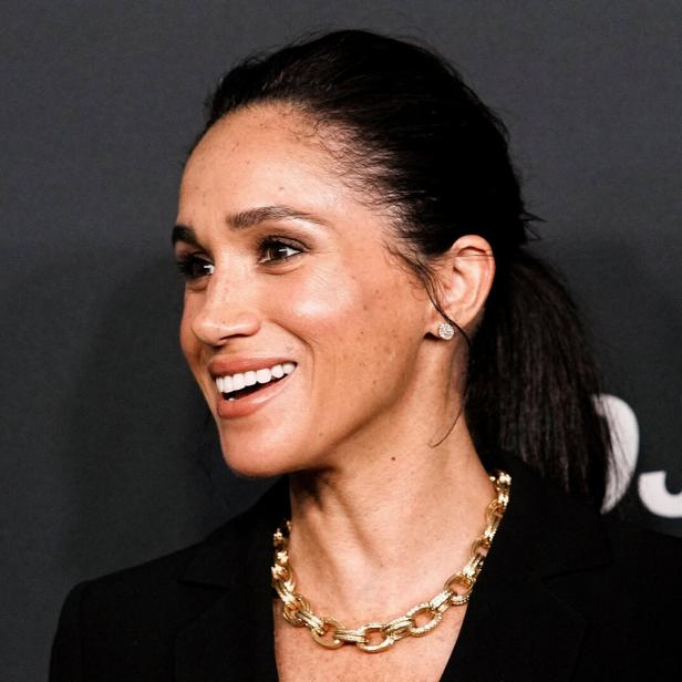 Herzogin Meghan lächelt mit goldenem Collier und Ohrring vor dunklem Hintergrund.