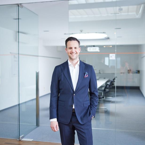 Ein Mann im Anzug steht lächelnd in einem modernen, hellen Büro mit Glastüren und Konferenztisch.
