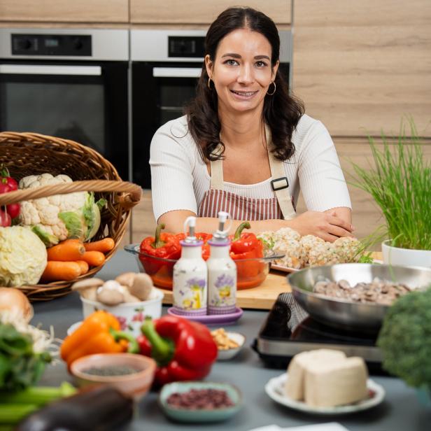 Frau in Küche mit vielen frischen Gemüsesorten und Kräutern auf dem Tisch.