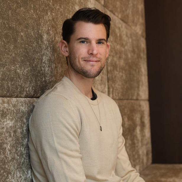 Ein junger Mann mit kurzem dunklem Haar sitzt entspannt vor einer gepolsterten, beigen Wand und lächelt leicht.