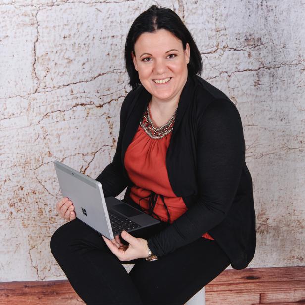 Frau mit dunklen Haaren sitzt lächelnd mit einem Laptop vor einer hellen Wand.