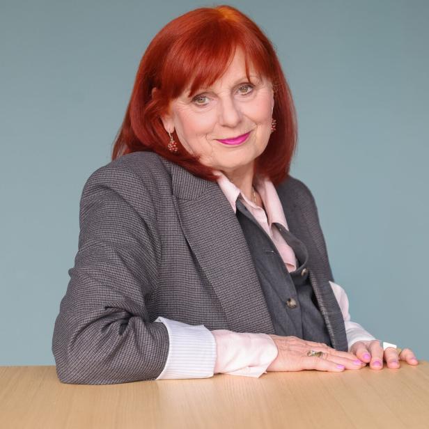 Eine Frau (Nora Frey) mit roten Haaren, Blazer und rosa Lippenstift lächelt in die Kamera.