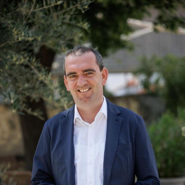 Ein Mann im blauen Anzug und weißem Hemd steht lächelnd im Freien vor grünen Pflanzen.