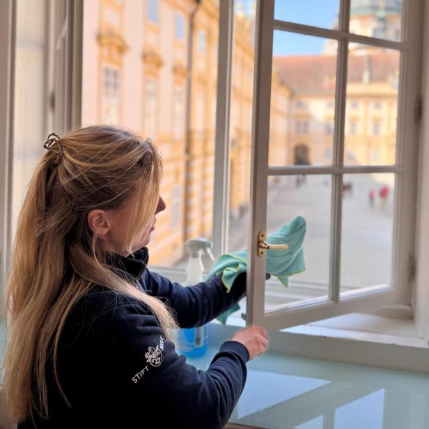 Frau reinigt ein geöffnetes Fenster im Stift Melk mit Blick auf den barocken Innenhof.