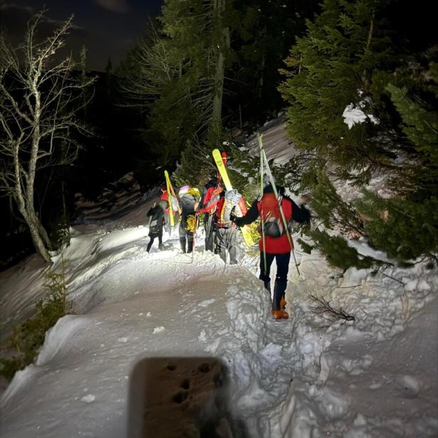 Mehrere Personen wandern nachts mit Skiern auf verschneitem Waldweg bergauf.