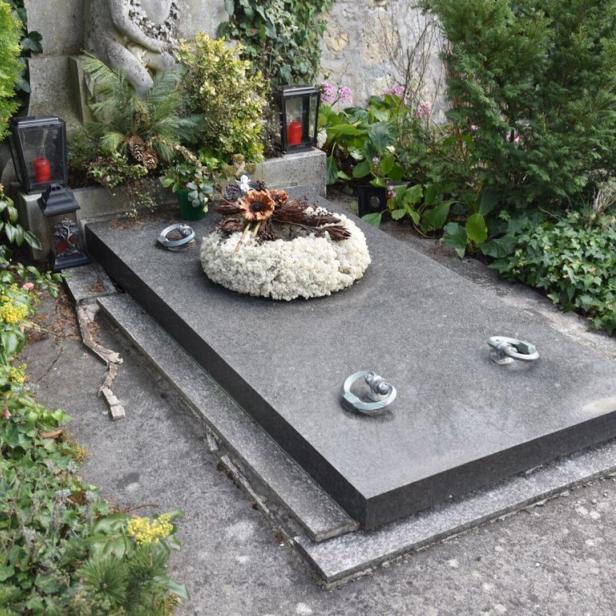 Grabstein mit Kranz aus weißen Blumen, drei kleinen Skulpturen und umgeben von grünen Pflanzen und Laternen mit roten Kerzen.