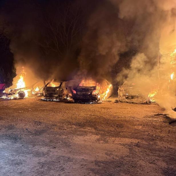 Mehrere Autos stehen in Flammen, dichter Rauch steigt in die Nacht auf, im Vordergrund ein Schotterplatz.