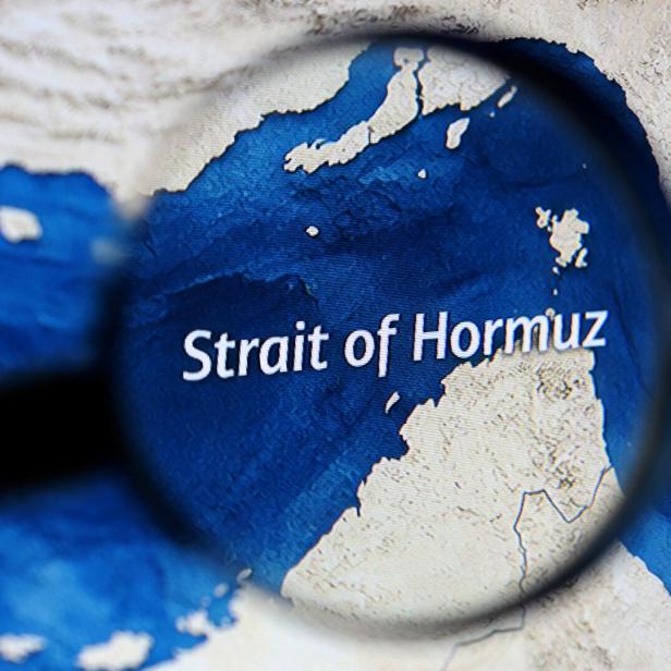 Eine Lupe vergrößert den Schriftzug „Strait of Hormuz“ auf einer Landkarte mit blauem Meer und beigen Küsten.